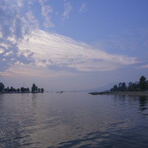 View from Sunset Beach, over English Bay. The sky is mostly blue with some small clouds, and the water is still. There is greyish haze on the horizon, partially hiding the mountains