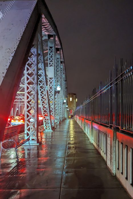 Burrard Bridge sidewalk at night. The pavement is wet, reflecting the red of taillights