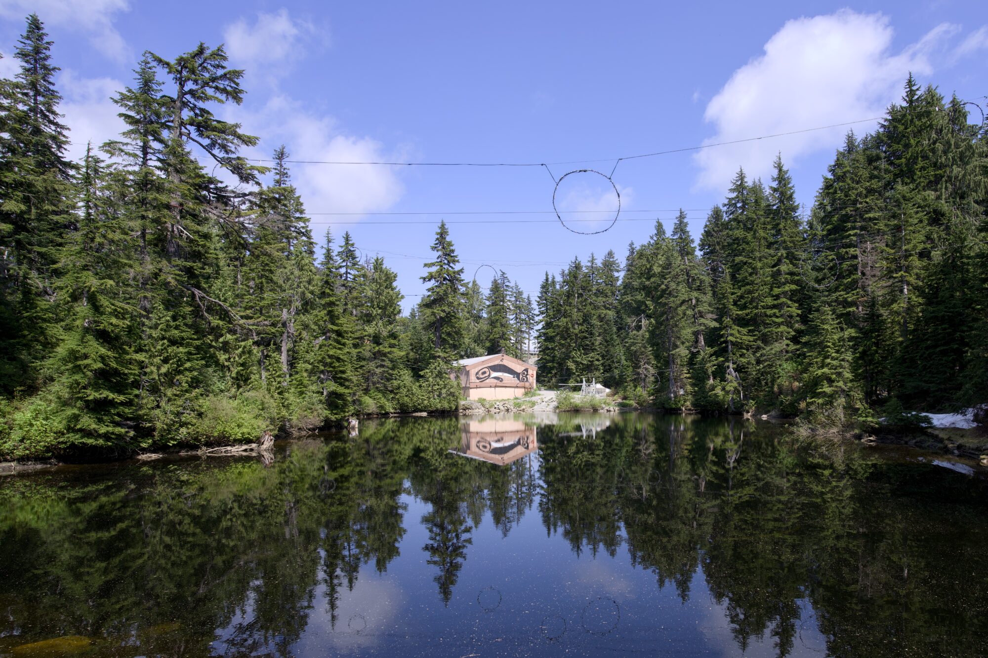 A little lake in the mountains, under blue sky and tall conifers. There are wires overhead, and a wooden structure with an Indigenous-style orca painted on, on the other side