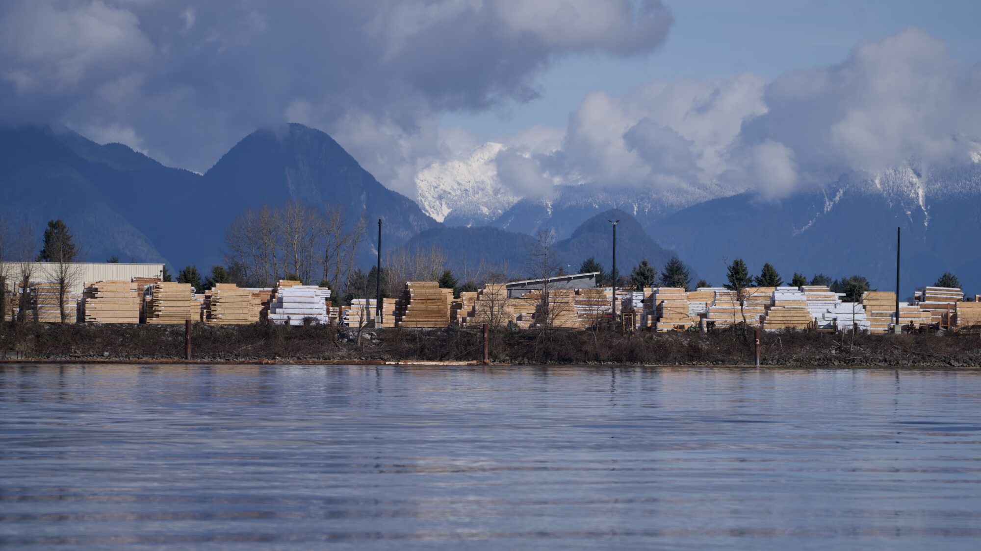 A view of planks and containers and assorted stuff across the river, and in the background some blue mountains mixed with clouds