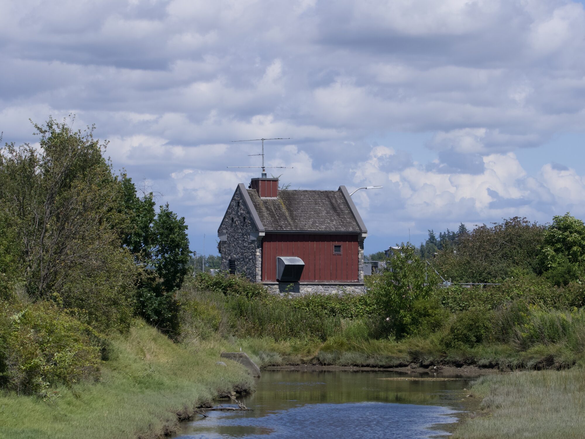 The Maple Street Pumphouse, a small building with a grey stone side and red side facing us. It is at the end of a little creek. Sky above it is mostly cloudy