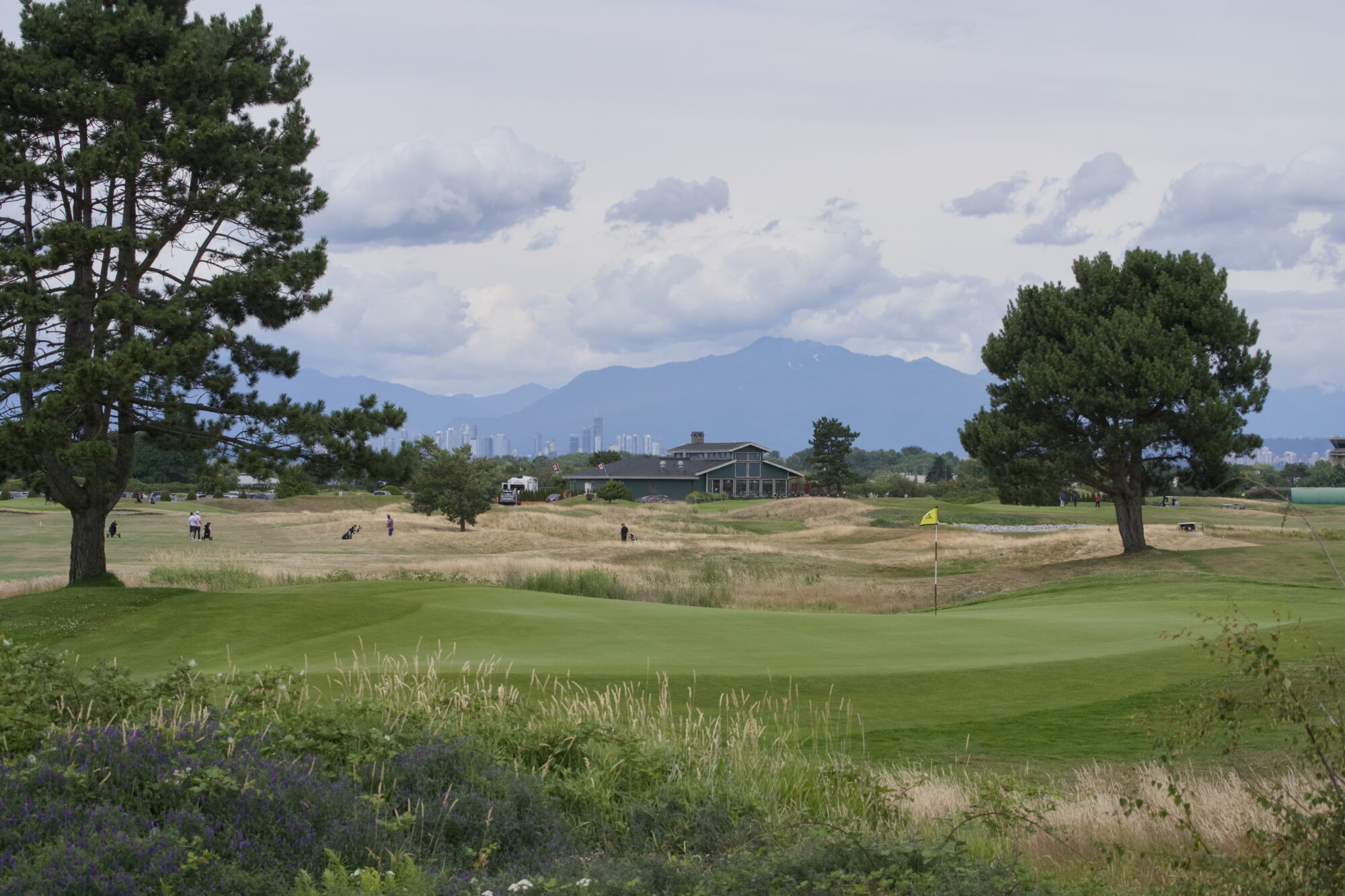 A gold course, with a few golfers out and about. In the background is the club house, and behind that are hazy blue mountains with some clouds