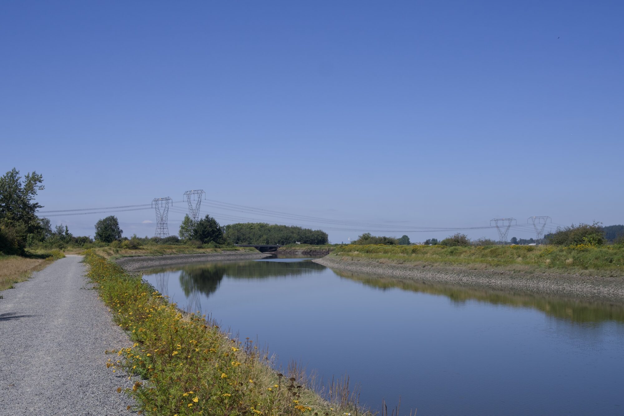 The Serpentine River, with a gravelly trail along one side and power lines in the distance. The water is still, reflecting the blue sky