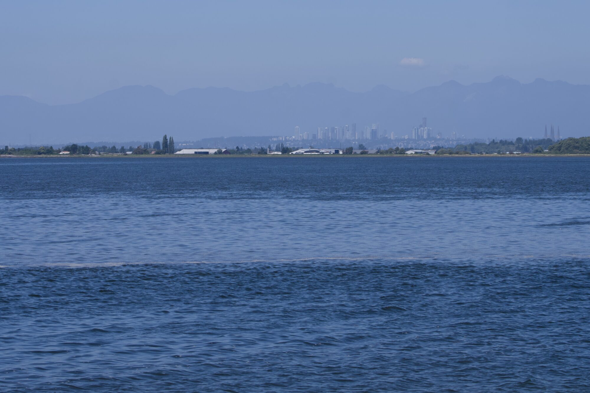 View from Blackie Spit: Blue water, some buildings on the opposite shore. Behind that, towers and residential neighbourhoods. Behind that, hazy blue mountains