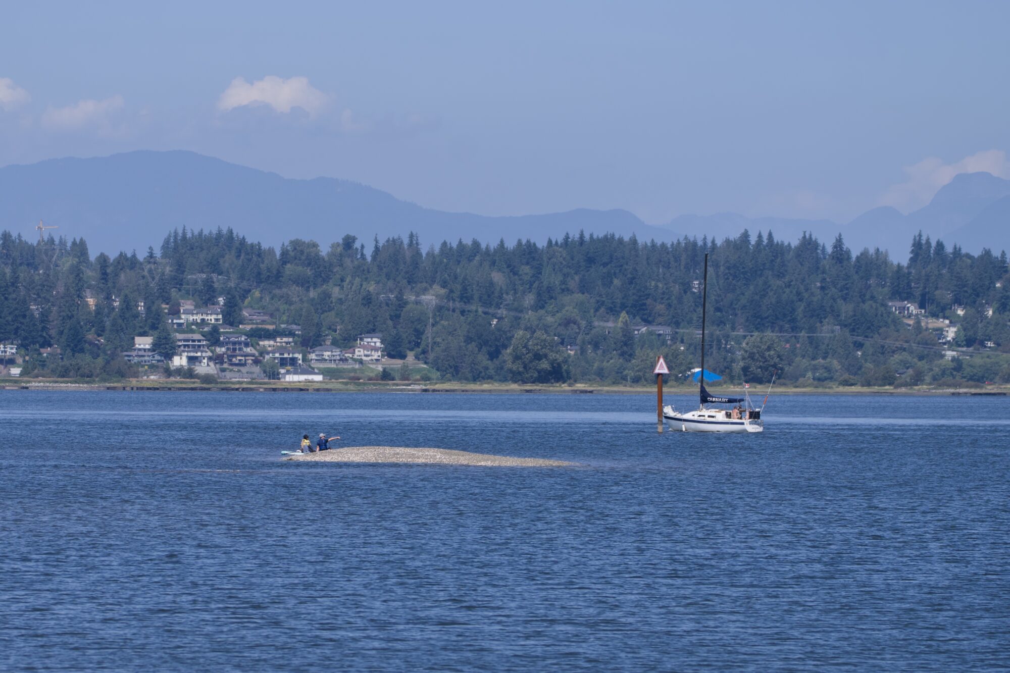 A couple of people sitting down on a sandbank out on the water. There is a small boat passing by. On the opposite shore are a few big houses surrounded by trees. Behind that, hazy blue mountains